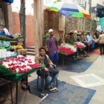 Amidst vibrant stalls and fragrant blooms, the marketplace near Nizamuddin sets the stage for the enchanting Thursday Poetry Nights at Nizamuddin. From the following image description, write the following incorporating the focus keyphrase seamlessly: Alt Text: Title: Caption: Description: Content supplied: Focus Keyphrase: Thursday Poetry Nights at Nizamuddin Description of the image: A young man sits at a flower stall in a bustling marketplace, surrounded by other vendors and shoppers. The image shows a narrow alleyway, possibly in India, with a stone wall on one side and various vendors' stalls on the other. The main focus is a young man with dark hair and a light brown jacket, sitting on a low stool behind a green-covered table laden with stacks of white containers, possibly for offerings, and an abundance of red and white flowers neatly arranged. He appears calm and relaxed. Behind him are several more stalls, each displaying similar items: more flowers, prayer mats (green with what appears to be Arabic script), and other small goods. Behind these stalls are several men sitting, some appearing to be vendors themselves, casually engaging in what seems to be commercial activity. They are dressed in varying styles of traditional clothing. A brightly colored umbrella stands prominently in the background, providing shade. In the background, a further group of people walk down the alley, a mix of men and women in various clothing suggesting a lively market atmosphere. The mood is one of calm industriousness, a peaceful bustling scene capturing daily life in a possibly religious context given the prayer mats and the apparent setting. The overall atmosphere is one of vibrant activity within a somewhat confined space, with a hint of warmth in the color palette and textures of the scene.